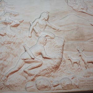 May include: A white ceramic relief sculpture depicting two Native American figures hunting deer in a forest setting. The sculpture is titled "Two Indians Stalking Deer" and signed "Norman Lungh".
