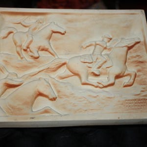 May include: A rectangular, off-white relief sculpture depicting a scene with three horses and riders. The riders are dressed in period clothing. The sculpture has a textured surface and is likely made of plaster or a similar material.