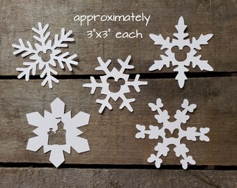 Puede incluir: Cinco copos de nieve de papel blanco, cada uno de aproximadamente 7,6 cm por 7,6 cm, con una silueta de Mickey Mouse recortada en el centro.