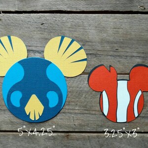 May include: Two paper cutouts of Mickey Mouse ears, one with a blue and yellow ocean-themed design, measuring 5 inches by 4.25 inches, and the other with an orange and white clownfish design, measuring 3.25 inches by 3 inches.