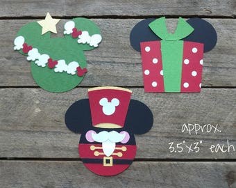 Puede incluir: Tres recortes de papel, cada uno de aproximadamente 8,9 cm por 8,9 cm. El primer recorte es un árbol de Navidad verde con adornos rojos y blancos. El segundo recorte es un regalo rojo y verde con lunares blancos. El tercer recorte es un Mickey Mouse rojo y negro vestido de tamborilero.