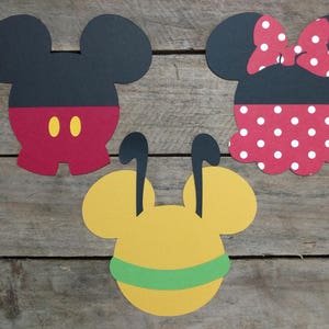 May include: Three paper cutouts of cartoon characters: Mickey Mouse, Minnie Mouse, and a yellow character with large ears. Mickey Mouse is wearing red shorts with black buttons and black shoes. Minnie Mouse is wearing a red polka dot dress with a red bow. The yellow character is wearing a green collar.