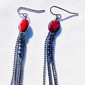 Può includere: Un paio di orecchini pendenti lunghi con una pietra preziosa rossa e più catene nere.