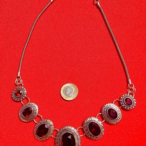 Op de afbeelding: Zilverkleurige ketting met ovale, donkerrode edelstenen. De ketting heeft een ketting en decoratieve zettingen rond de stenen. De achtergrond is effen rood.