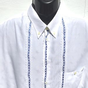 Puede incluir: Una camisa blanca de manga corta con botones y ribetes azules y blancos en la parte delantera.