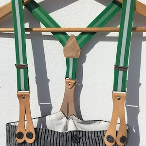 May include: Green and white striped suspenders with brown leather straps and metal clips. The suspenders are hanging on a wooden hanger.