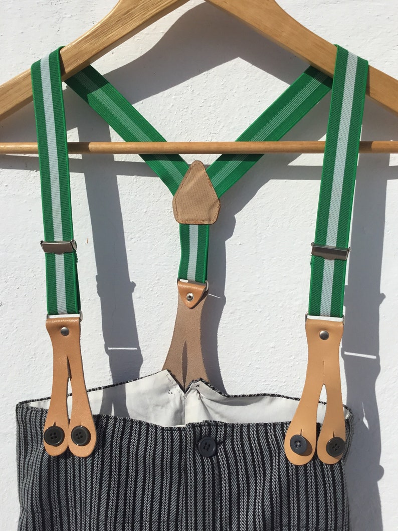 May include: Green and white striped suspenders with brown leather straps and metal clips. The suspenders are hanging on a wooden hanger.