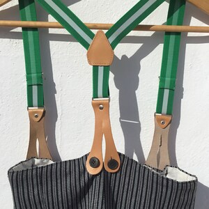 May include: Green suspenders with white stripes and brown leather accents. The suspenders are hanging on a wooden hanger and have two buttons on each side.