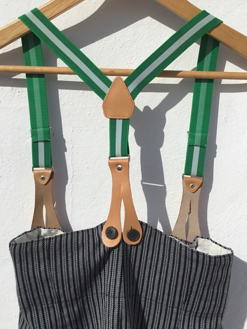 May include: Green suspenders with white stripes and brown leather accents. The suspenders are hanging on a wooden hanger and have two buttons on each side.