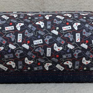 May include: A rectangular, zippered bag with a dark blue background covered in a pattern of white, gray, and red video game controllers and consoles. The bag has a dark blue base and gray zipper pulls with a game controller charm.