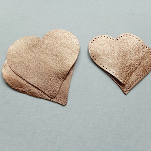 Può includere: Due pezzi di pelle a forma di cuore di colore oro rosa. Il cuore più grande è sovrapposto a un cuore più piccolo.