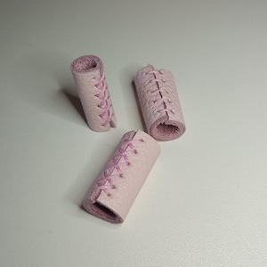May include: Three pink leather cord ends with pink stitching. The cord ends are cylindrical and have a smooth finish.