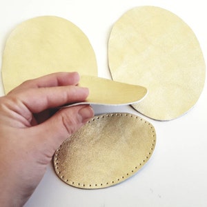 Può includere: Tre forme ovali in pelle dorata, una è tenuta da una mano. La pelle è cucita sui bordi.