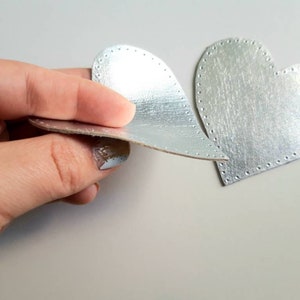 May include: Two silver heart-shaped cutouts. One is held by a hand, showing a layered construction. The other heart lies flat, with a stitched edge. The hearts have a textured surface, possibly craft supplies for a DIY project.