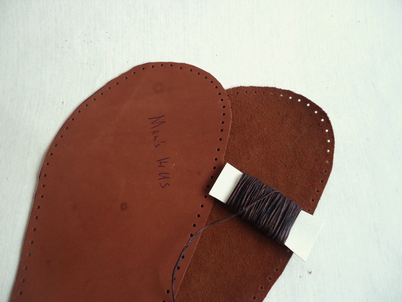 May include: Two brown leather pieces with holes around the edges, one with the text "Men's 14 US" written on it. A spool of brown thread is on top of the leather pieces.