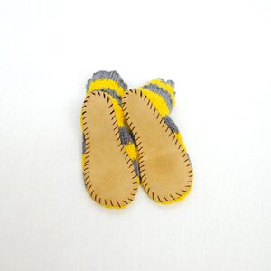 May include: A pair of gray and yellow striped knitted slippers with leather soles. The slippers have a stitched seam around the edge of the leather soles.