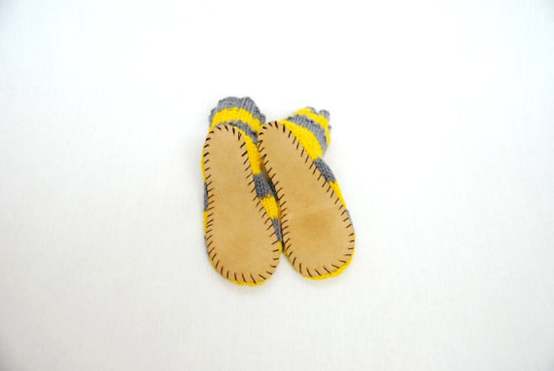 May include: A pair of gray and yellow striped knitted slippers with leather soles. The slippers have a stitched seam around the edge of the leather soles.