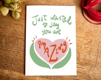 May include: A white greeting card with the words "Just wanted to say you are AMAZING" in green and orange. The word "AMAZING" is inside a pink heart shape, with green leaves around it. The card is on a wooden surface.