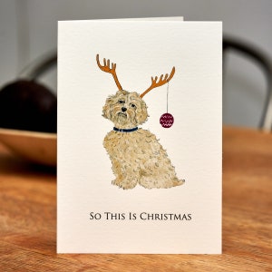 Pode incluir: Um cartão de Natal com uma ilustração em aquarela de um cão usando chifres de rena e uma bola vermelha. O texto "SO THIS IS CHRISTMAS" está impresso abaixo do cão.
