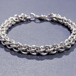 May include: A silver chain bracelet with a delicate, intricate design. The bracelet is made of small, linked rings that create a woven pattern.