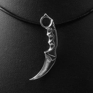 Puede incluir: Un colgante de cuchillo karambit plateado en un cordón de cuero negro. El cuchillo tiene una hoja curva y un anillo para el dedo.