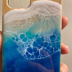 May include: A blue and white resin phone case with a wave design. The case is made of wood and has a clear resin coating. The wave design is made with white and blue resin and has a glittery finish.