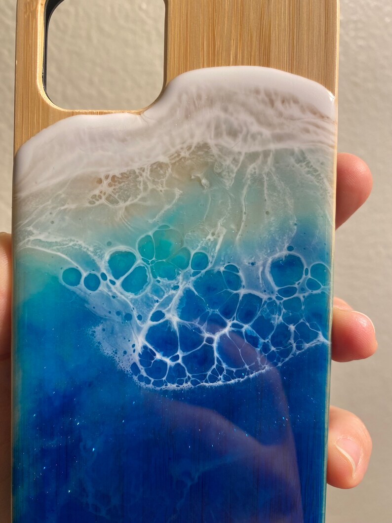 May include: A blue and white resin phone case with a wave design. The case is made of wood and has a clear resin coating. The wave design is made with white and blue resin and has a glittery finish.