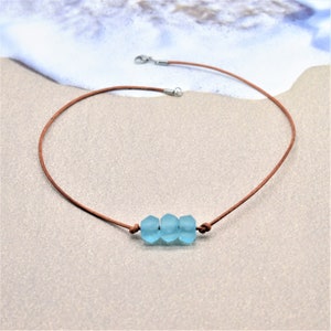 May include: A brown leather cord necklace with three light blue, faceted beads. The necklace has a simple design with a clasp closure. The beads are centered on the cord, creating a minimalist style. The necklace is displayed on a sandy surface.