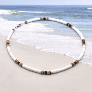 May include: A white, brown, and black beaded necklace with a silver clasp. The necklace is made of small, round beads and is lying on a sandy beach.