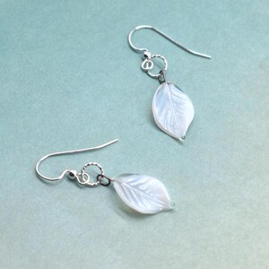 May include: A pair of delicate earrings featuring leaf-shaped pendants. The leaves are a translucent white with subtle veining, suspended from silver-colored hooks. The earrings are displayed against a soft, muted teal background.