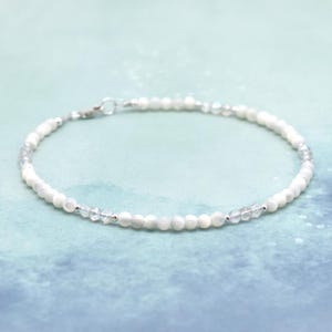 May include: A delicate white and silver beaded bracelet with a silver clasp. The bracelet is made with small, round beads and has a subtle shimmer.