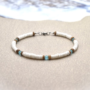 May include: White shell bracelet with wooden and blue accents. The bracelet is made of small, flat white shells, interspersed with small wooden beads and light blue beads. The bracelet is on a sandy surface.