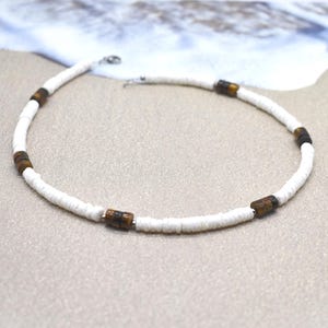 May include: White beaded necklace with brown cylindrical accents. The necklace is made of small, round white beads and features several dark brown, barrel-shaped beads. The necklace is displayed on a sandy surface.