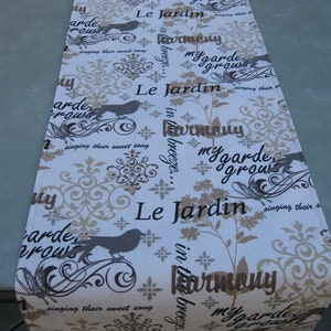 May include: A white table runner with a beige and brown floral pattern. The text "Le Jardin" is repeated throughout the design, along with the phrases "in the breeze... harmony" and "my garden grows".  The runner features a repeating pattern of stylized birds and floral motifs.