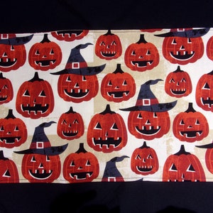 May include: A Halloween-themed fabric with a repeating pattern of orange jack-o'-lanterns wearing black witch hats. The pumpkins have carved faces and are set against a beige and white background. The fabric is likely used for seasonal decor.