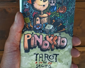 Może przedstawiać: Prostokątna karta tarota z kolorową ilustracją drewnianej marionetki z długim nosem, otoczonej kwiatami, sercami i czaszkami. Tekst głosi PINOKIO TAROT 11 by 20 SAGITTIS czarnymi i pomarańczowymi literami.