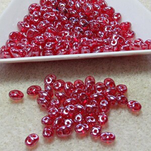 May include: A collection of small, translucent red glass beads, each featuring a distinct two-hole design. The beads have a smooth, glossy finish and are presented both inside a white triangular tray and scattered loosely on a textured surface.