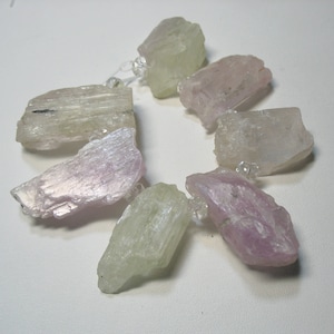 May include: A necklace featuring rough-cut gemstones in shades of pale pink, green, and clear. The stones are strung together with small, clear beads. The gemstones have a natural, textured appearance, and are arranged in a circular pattern.
