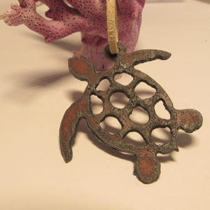 May include: A cutout metal turtle pendant with a brown and red patina. The turtle has a detailed shell with a cutout design. The pendant is hanging from a brown leather cord.