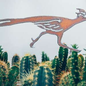 May include: A metal roadrunner silhouette in a running pose, with a rusty brown finish. The bird is set against a backdrop of green cacti with sharp spines. The roadrunner is a symbol of speed and the American Southwest.