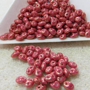May include: A collection of small, oval-shaped glass beads with a metallic pink finish. Each bead features two distinct holes positioned side-by-side. The beads have a smooth, rounded surface with a reflective sheen, displayed in a pile.
