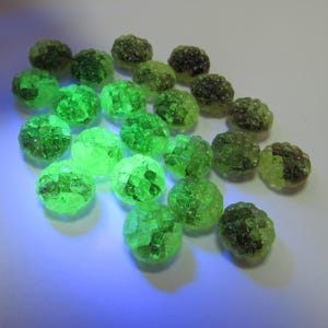 May include: A collection of green, spherical beads with a textured surface. The beads vary in shade from a bright, almost neon green to a darker, olive green. The arrangement is scattered across a white surface.