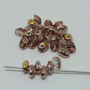 May include: A pile of small, clear glass beads with a rose gold iridescent finish. The beads are oval shaped and have a small hole in the center. Some of the beads have a gold accent around the hole. A few beads are strung on a thin wire.