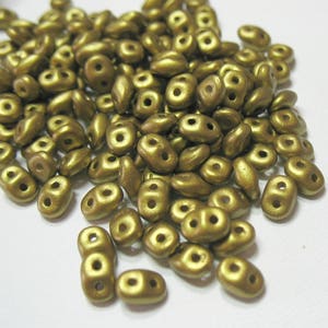 May include: A pile of small, gold-colored glass beads with two holes each. The beads are shaped like a teardrop with a rounded top and a pointed bottom.