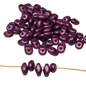 May include: A pile of small, dark purple, metallic, glass beads. The beads are shaped like a teardrop with a hole in the center. A strand of the beads is strung on a gold wire.