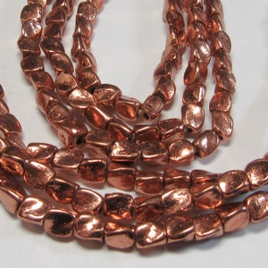 May include: A strand of copper-colored beads with a hammered, irregular texture. The beads are oval-shaped and have a metallic sheen.
