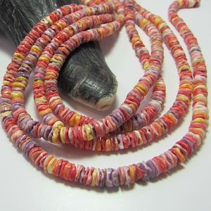 May include: Strands of colorful shell necklaces in shades of red, orange, yellow, and purple. The necklaces are made of small, flat shell pieces, creating a layered effect. The jewelry is displayed on a dark, textured surface.