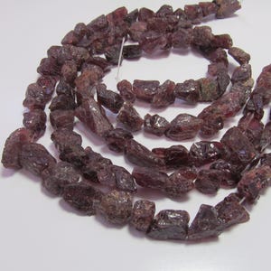 May include: Strand of rough-cut, dark red-brown garnet gemstone beads. The beads are irregularly shaped, with a natural, unpolished appearance. The gemstones are strung together on a white string, suitable for jewelry making.