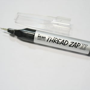 May include: A silver and black Thread Zap II tool with a clear plastic cap. The tool has a fine needle tip and the text "THREAD ZAP II" is printed on the silver body. The tool is designed for trimming threads.