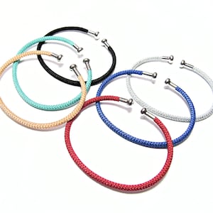 May include: A collection of open-ended bracelets in various colors, including black, turquoise, tan, red, blue, and silver. Each bracelet features a woven texture and silver-toned metal ends. The bracelets are arranged on a white background.